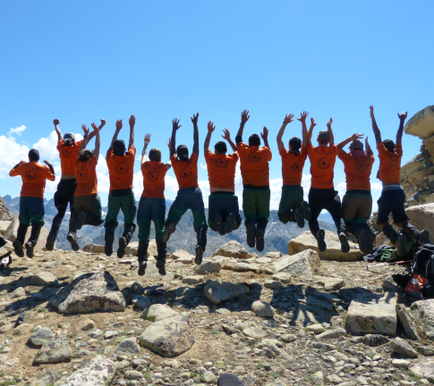 a group of people in orange shirts, facing away from the camera, are jumping in sync in mountainous terrain